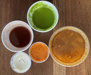 A colorful selection of chutneys and a tub of sambar soup accompany our takeout meal from Shreeji.
