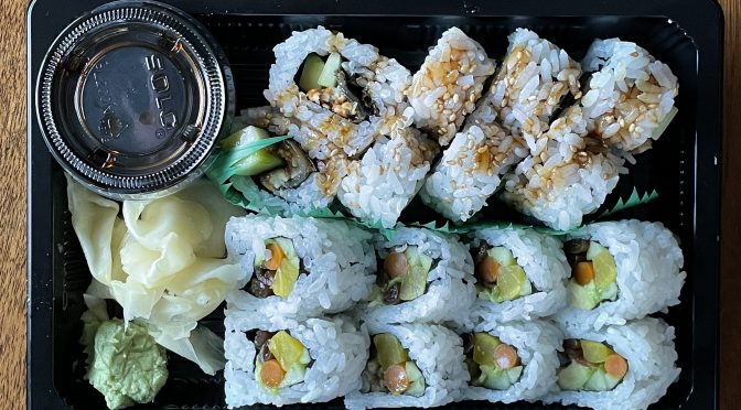 Two sushi rolls at hiko-A-mon: Eel roll at the top and veggie roll beneath, with pickled ginger, wasabi paste, and a tub of soy sauce on the side.