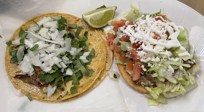 On the left, a pair of house-made corn tacos topped with falling-apart pork carnitas, chopped onion, and fresh cilantro. At right, sopesitos, a thick hand-made tortilla topped with lengua (beef tongue), lettuce, queso fresco, and crema.